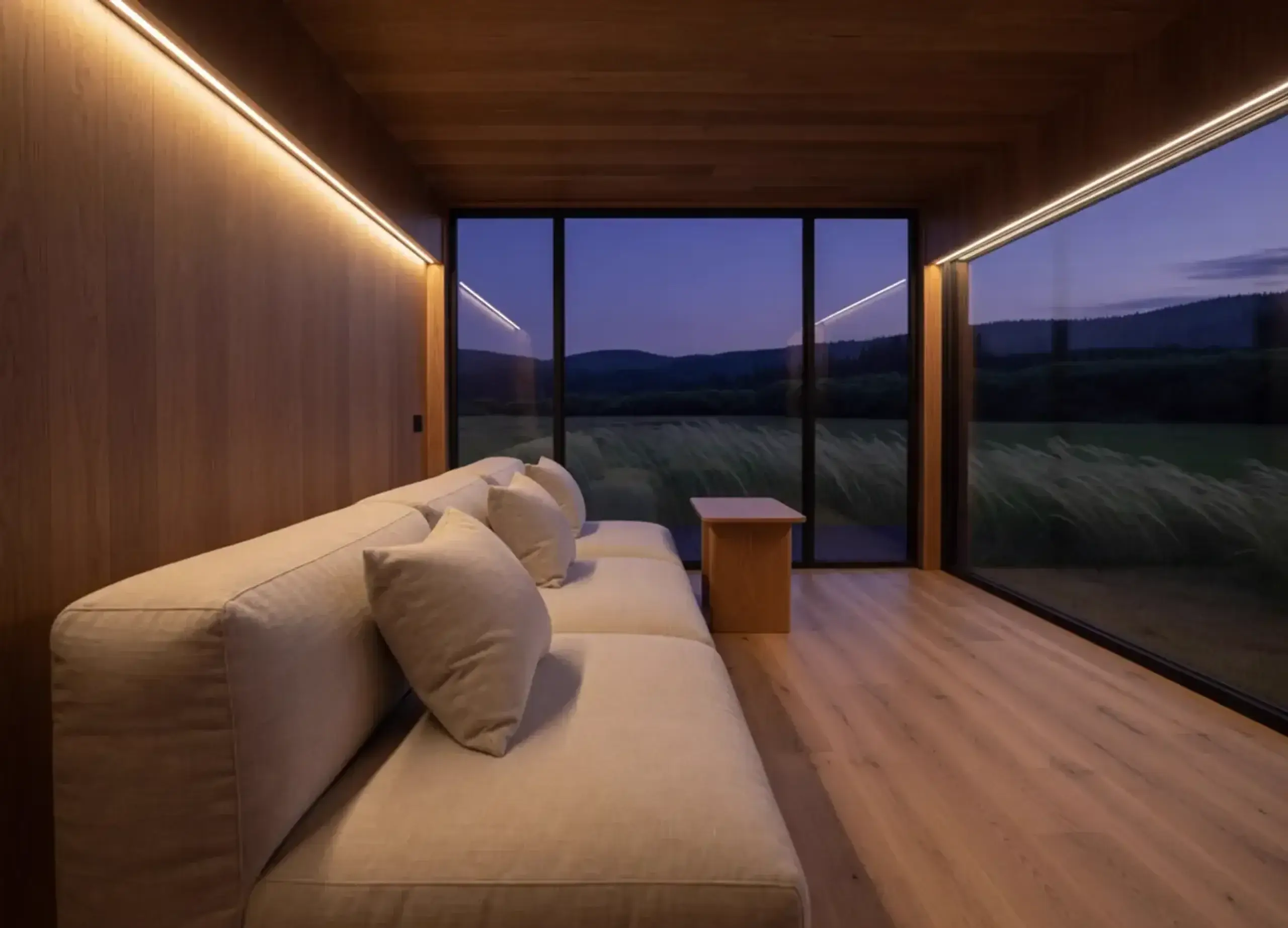 A linen sofa in a warm wood room, evening light along the ceiling, a meadow beyond the glass.